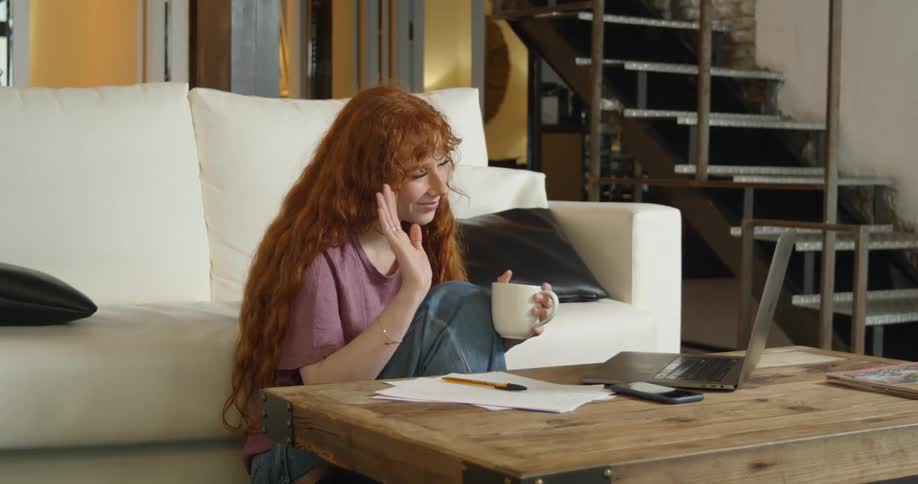 Redhead sitting on the floor of the living room talking on a video conference on a laptop