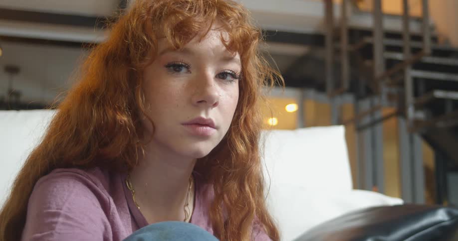 A young woman turns to look into the camera while sitting on a sofa indoors