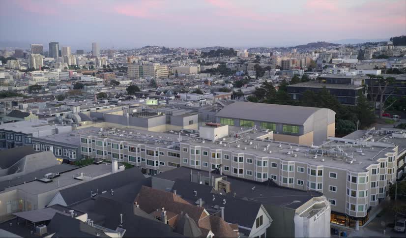An aerial view of cars houses and apartment buildings in a neighborhood with the downtown skyline in the distance
