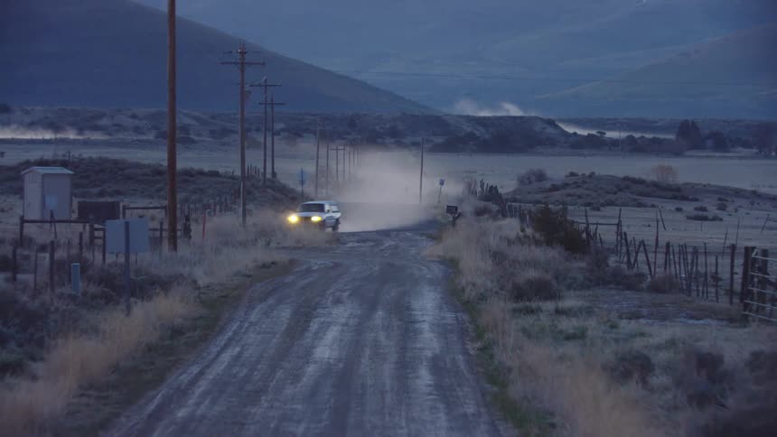 The camera travels down a dirt road and see's a white pickup truck with headlights on turn before them on a rural ranch deep in the mountains of Montana