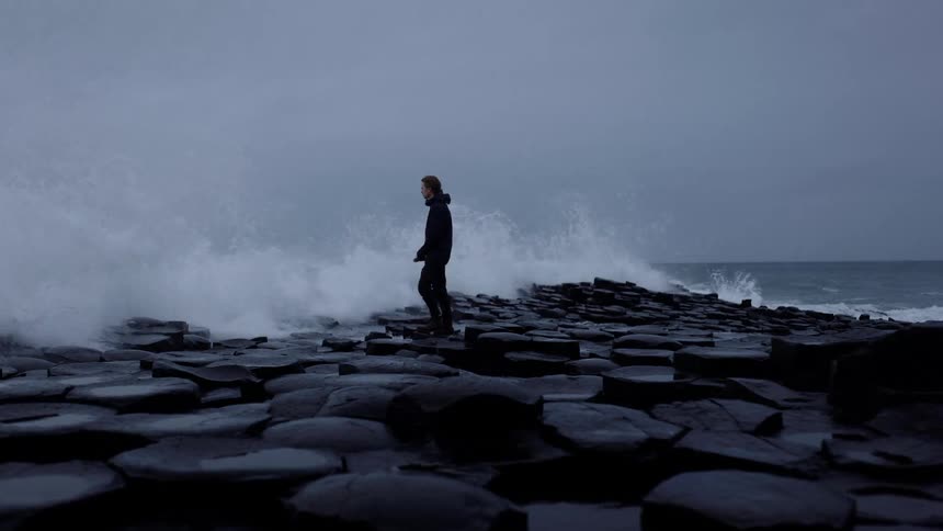 A man is splashed by ocean waves while standing on a rocky coastline on a cloudy day