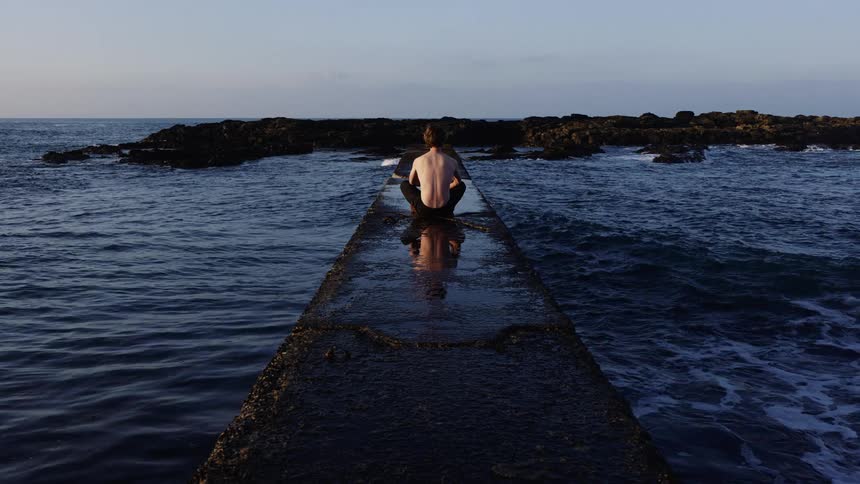 Shirtless man sitting on the concrete dock next to the ocean
