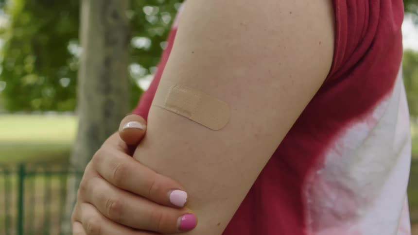 A woman standing outside with a band aid on her arm with a fence trees the sky and clouds in the background