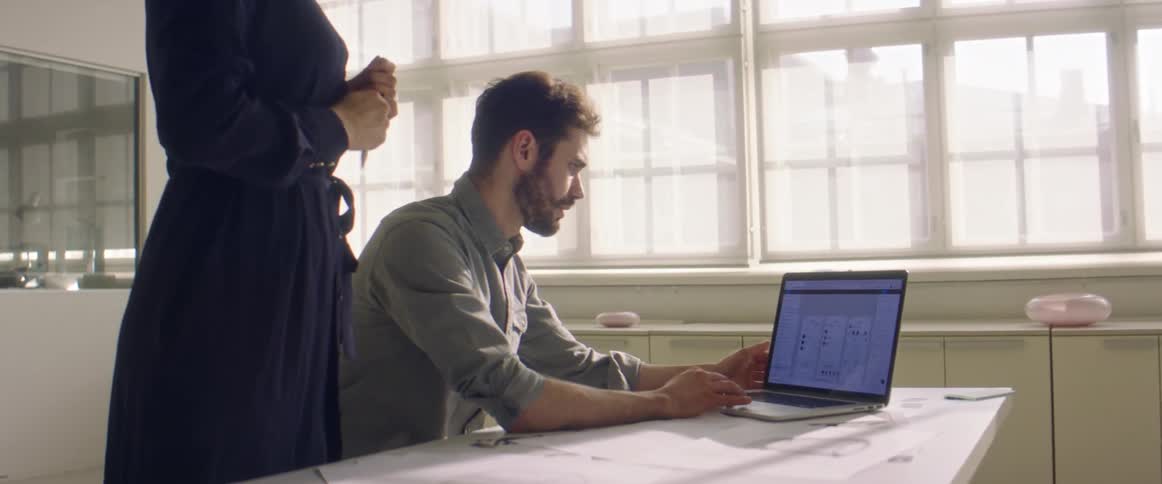 A man sits at a desk with a laptop and a woman talks behind him