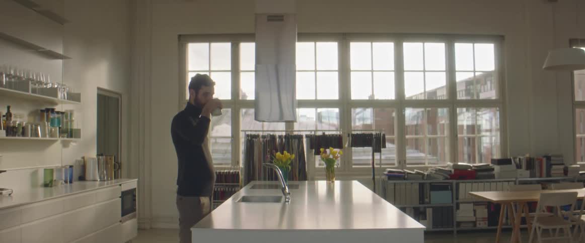 A man standing in a room in front of a sink and faucet and drinking from a cup