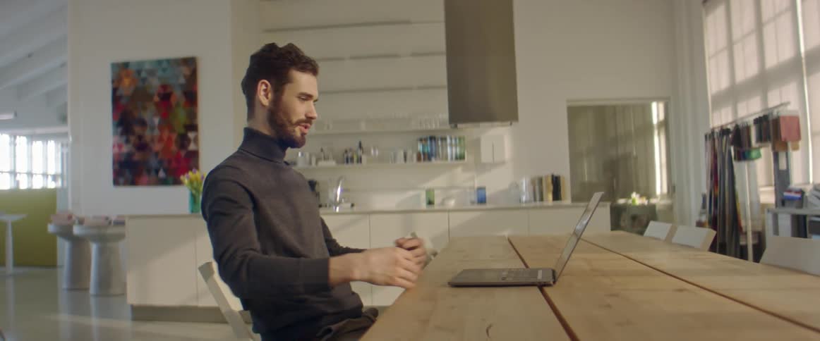 Man sitting at table in shop indicates his clothes while talking via video call on laptop