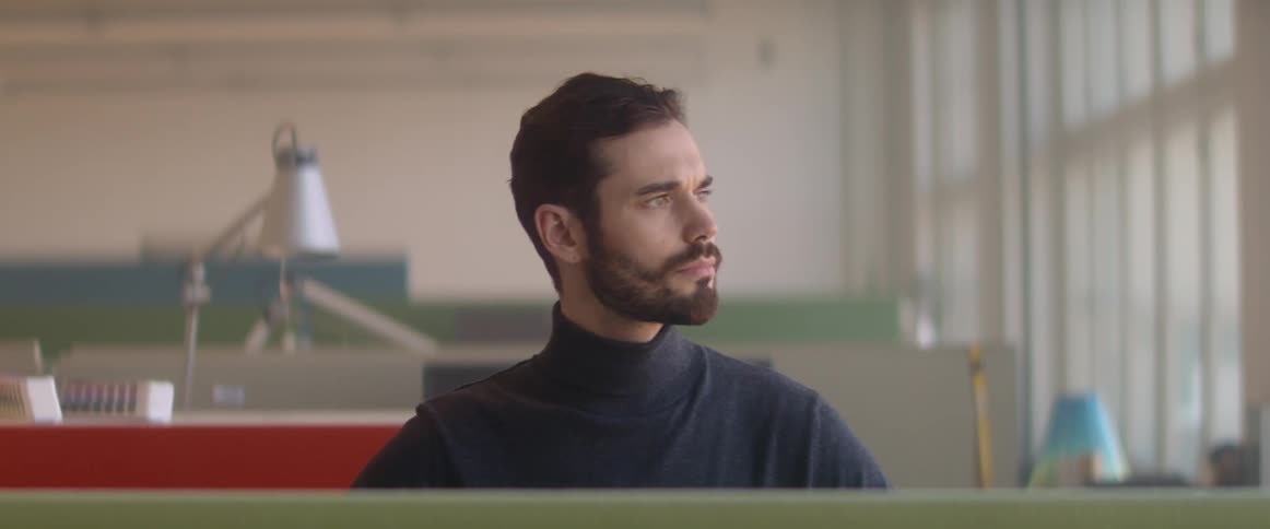 A man sitting at a desk in an office looks towards the window contemplatively for a moment before returning to his work