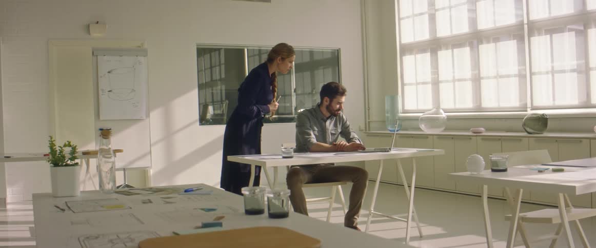 A man sits at a laptop with a woman looking over his shoulder and them talking about what they see in a large office room
