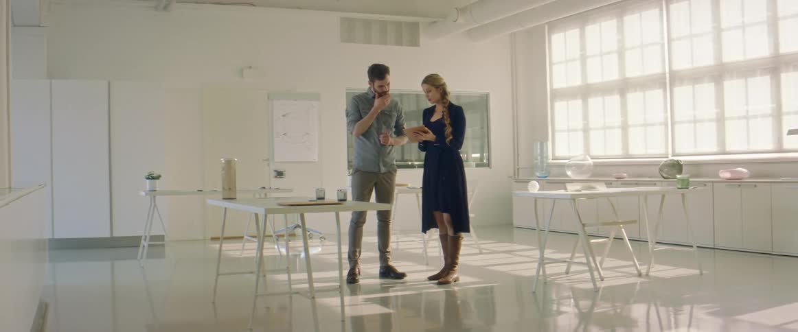 A man and a woman standing in a room with desk and chairs with a wall in the background
