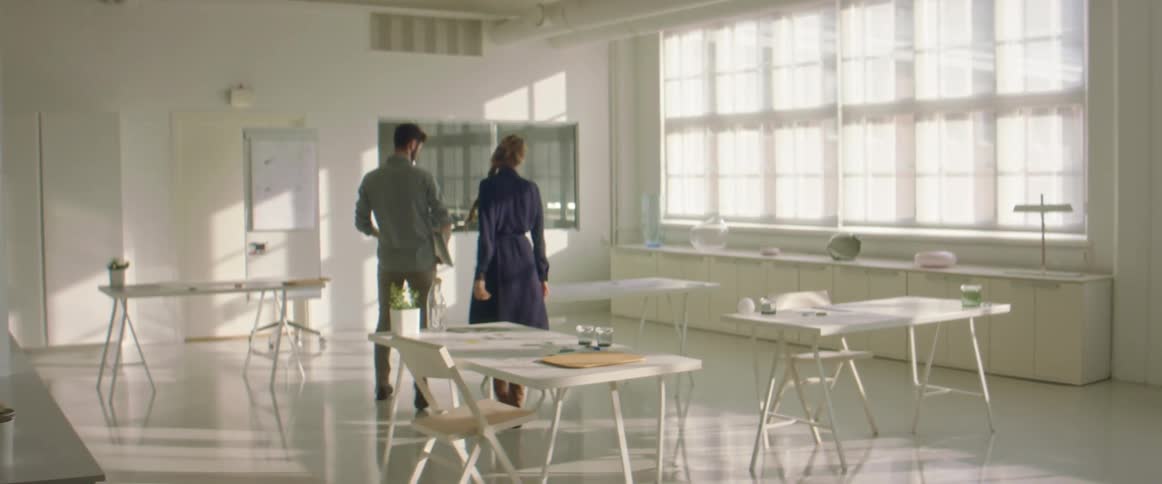 A man and woman walk into a large office space and sit down with a laptop
