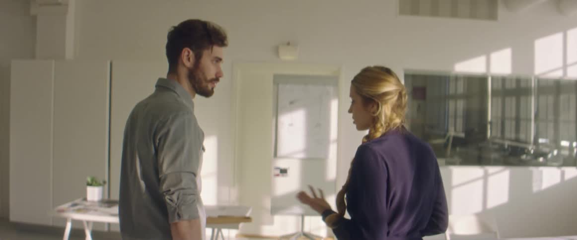 A man and woman walk in to an office and go to a desk where the man sits down and opens up his laptop