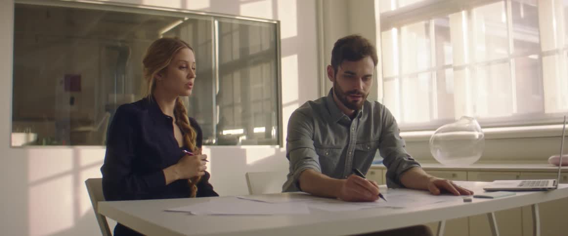 Blonde woman with a black sweater and man with a beard and a grey shirt sit at table and work together in a room