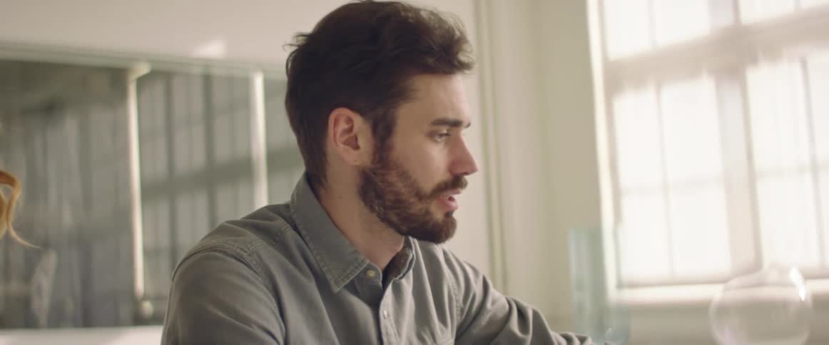 A man sits at a table looking at a laptop and speaking to the woman behind him