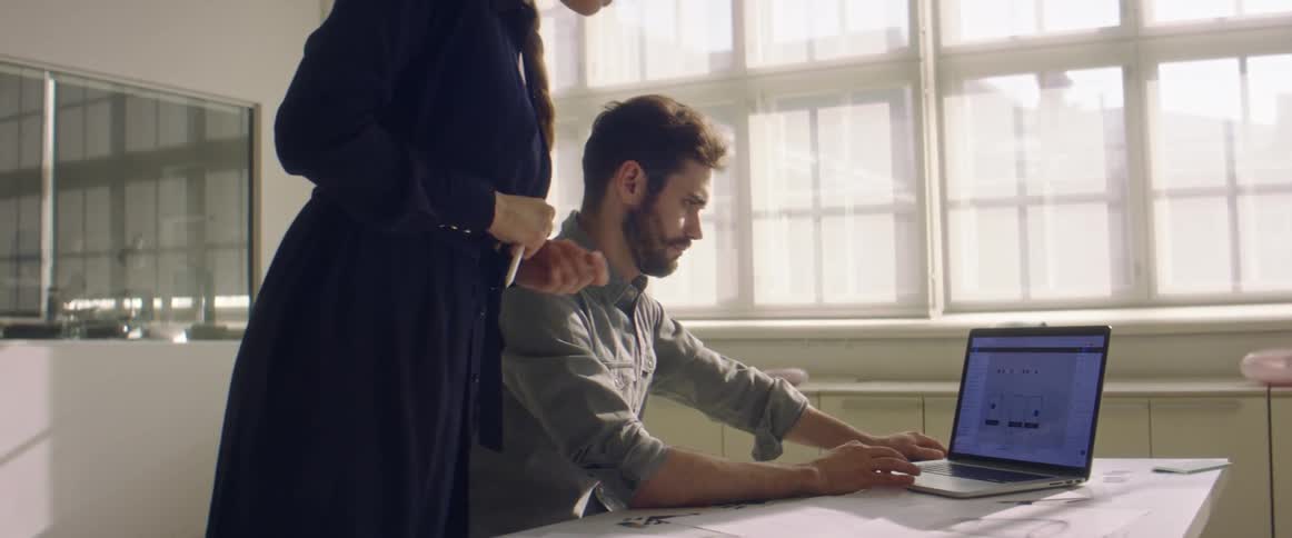 A man sits at a desk with a laptop and a woman talks to him from behind