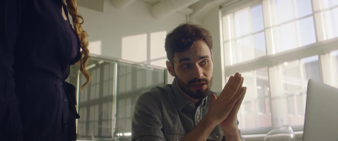 A man in a room sitting at a desk looking at a laptop with a wall and window in the background