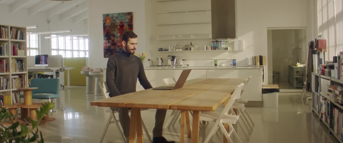 A man sits at a large table in an open floor plan office setting having a video call on a laptop