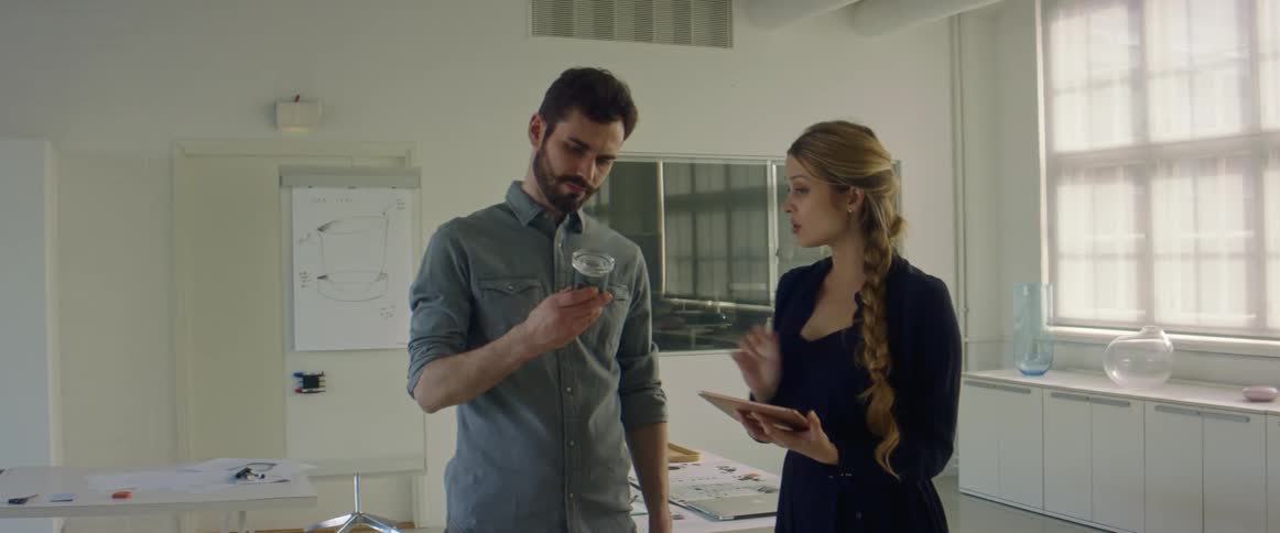 A man shows a small jar to a woman holding a tablet and stylus as they stand together in a conference room during the day