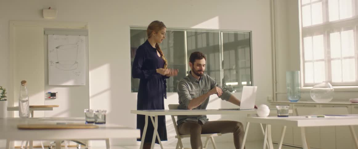 A woman speaks to a man sitting in front of a laptop at a desk indoors during the day