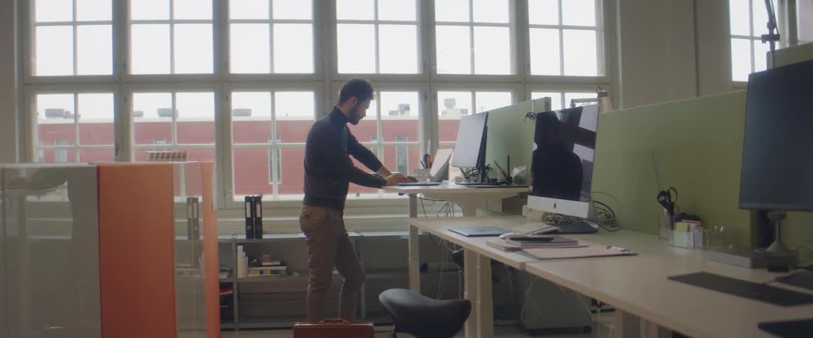 Bearded man stands at a desk and works on a laptop while an orange and white partition stands behind him