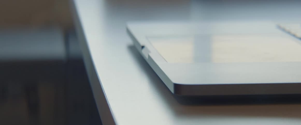 A person opens a laptop lying atop a table with one hand indoors during the day