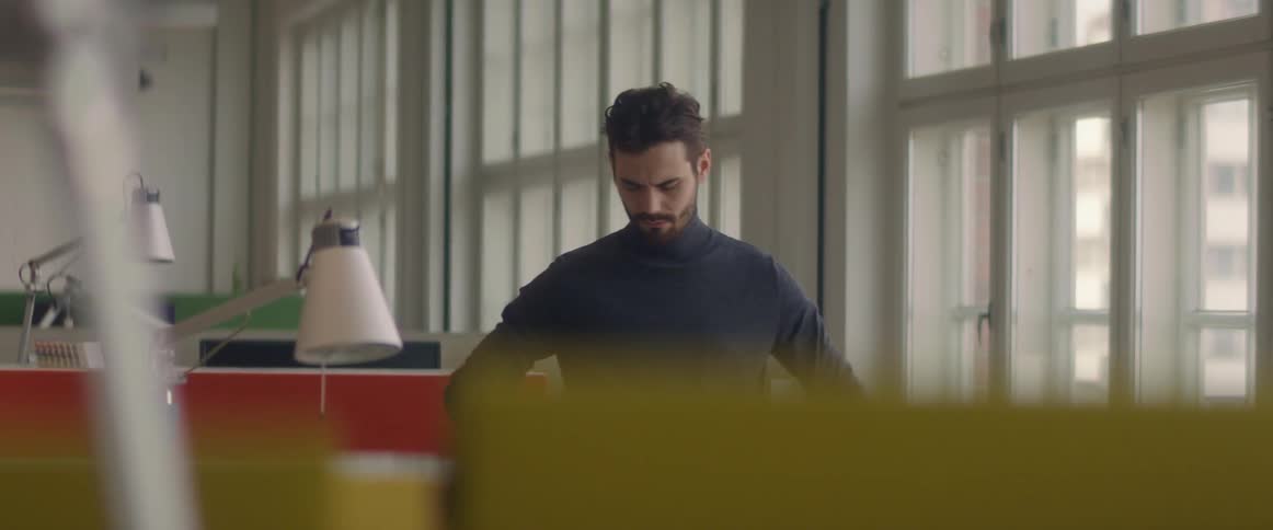 A man standing in an office strokes his beard contemplatively while looking between his desk and the window