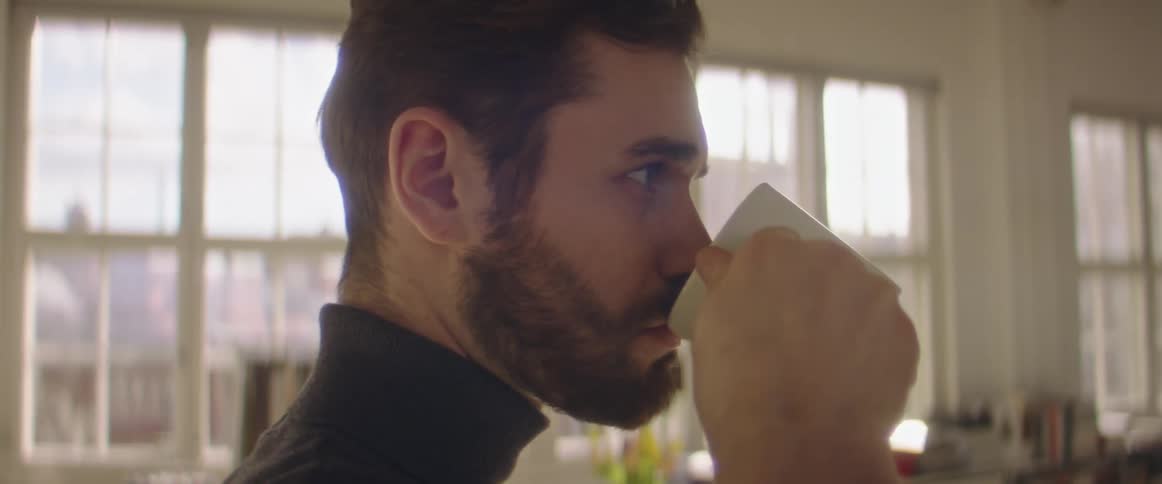 Man with a beard and a black turtleneck sweater sips coffee in a room