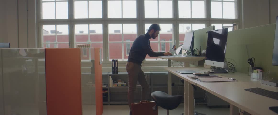 A man in a room standing by a desk and using a laptop with a wall and windows in the background