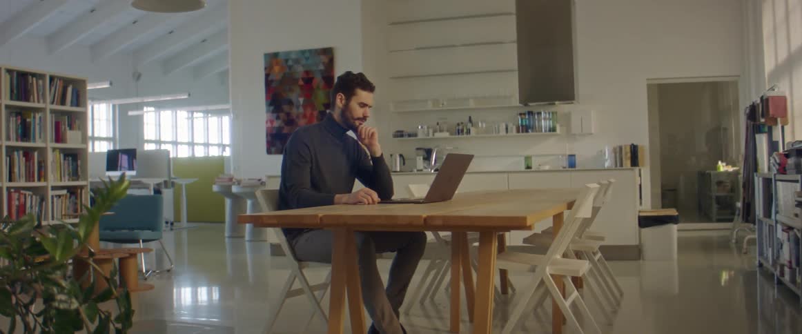 A man in a room sitting on a chair at a table using a laptop