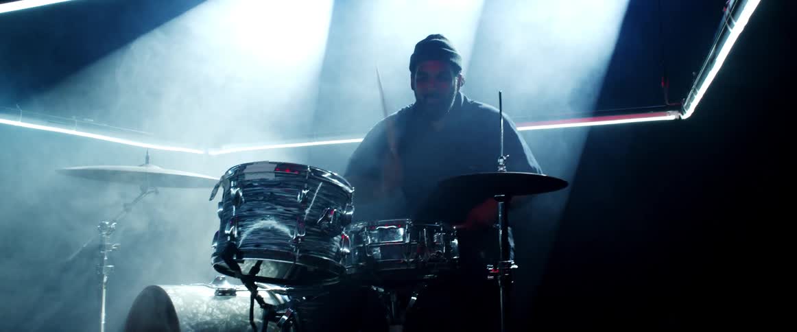 Woman with a beard and a beanie plays drums in a fog indoors