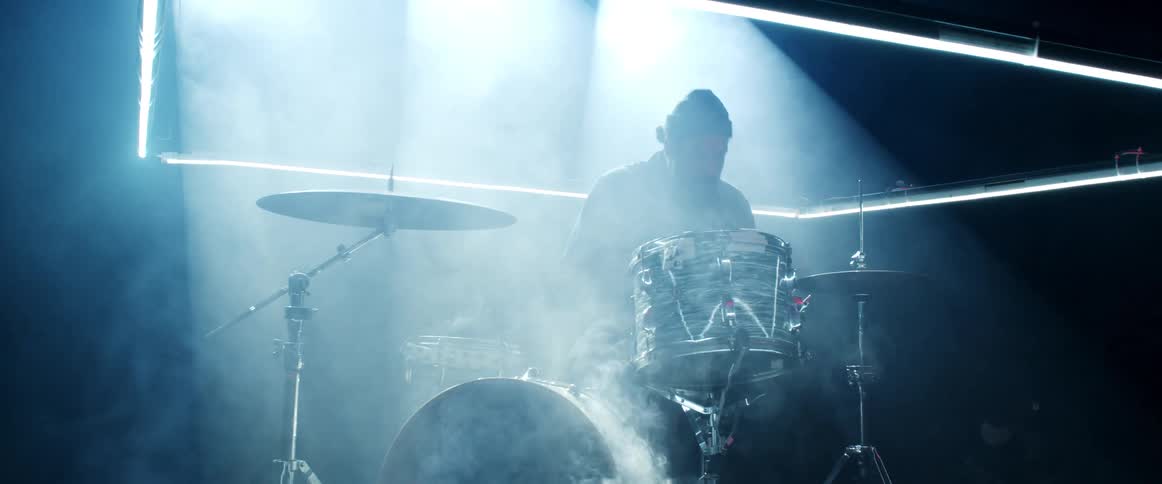 Man playing on a set of drums on a dark room surrounded by white smoke