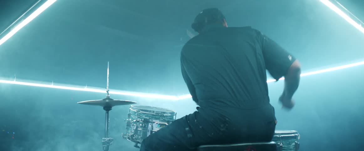 A man with a hat sitting in a room playing drums with drumsticks