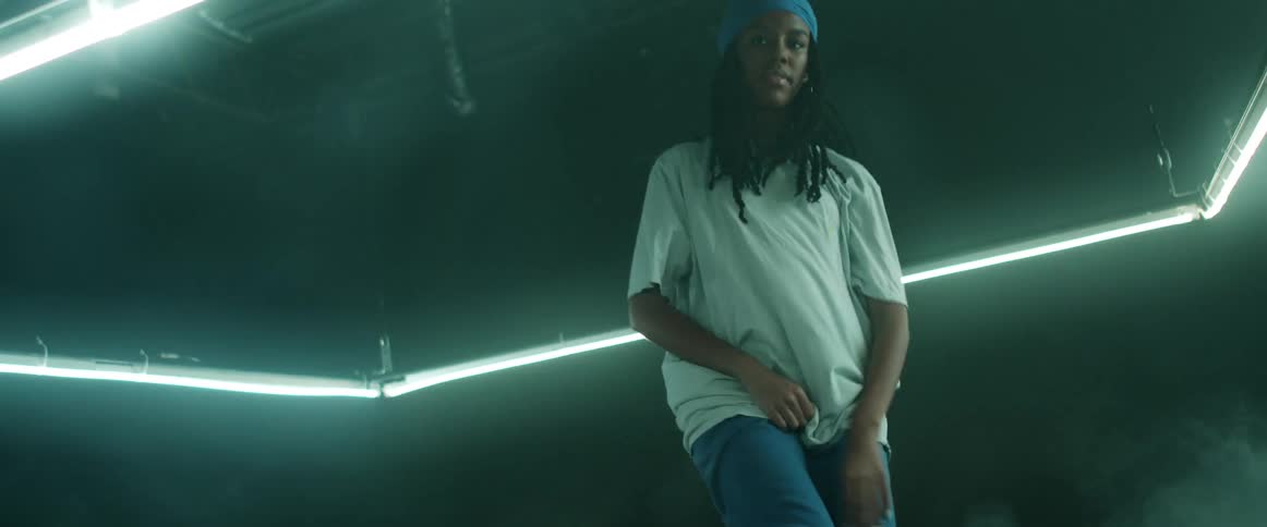 Woman with a blue headband braids hoop earrings and a white loose t shirt dances in a dark room with lights