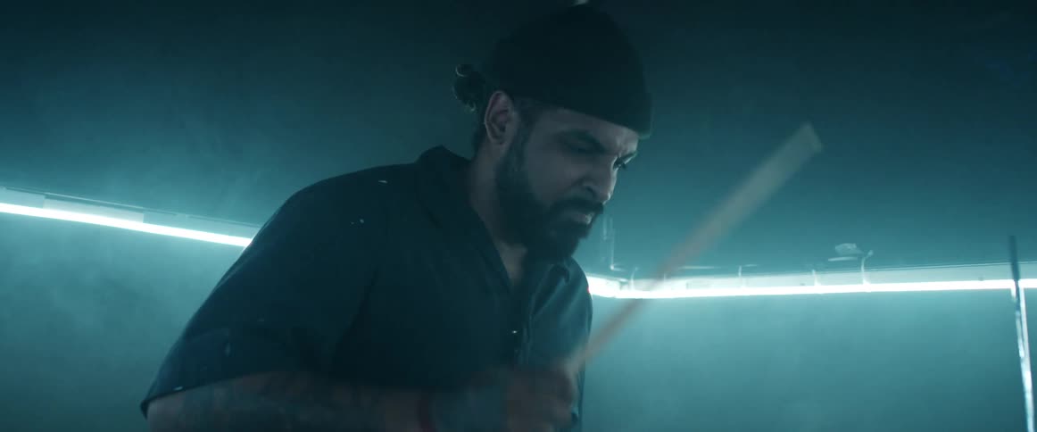 Man with beard wearing knit hat plays drums in room light by bright line lining the wall