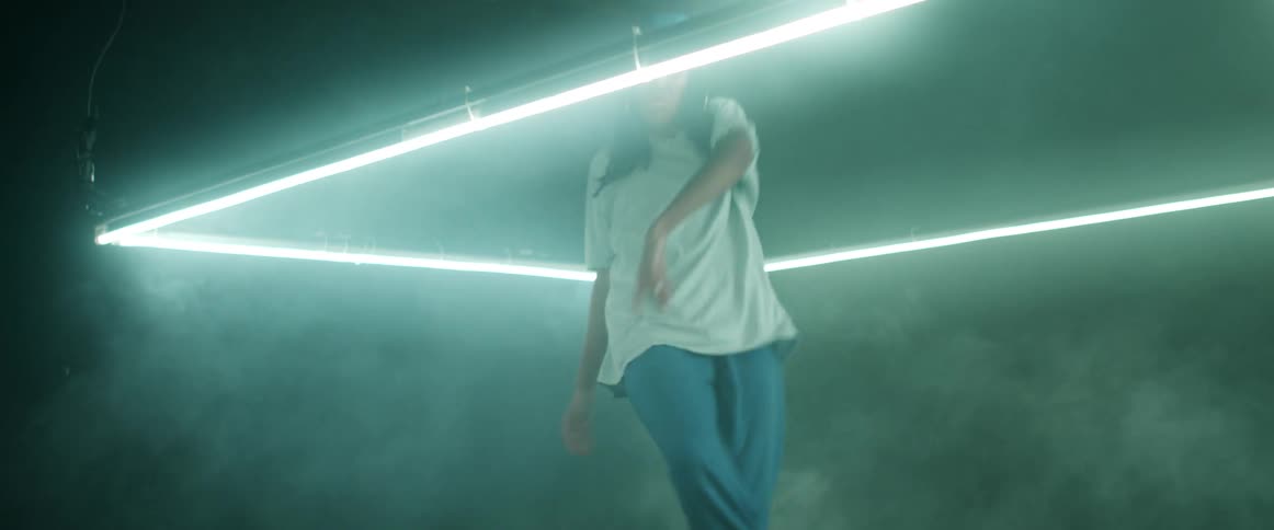 A woman in a backward hat dances hip hop inside a glowing frame that hangs from a ceiling