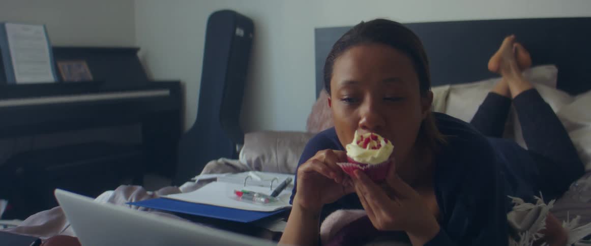 A woman laying in bed looking at a laptop and eating a cupcake