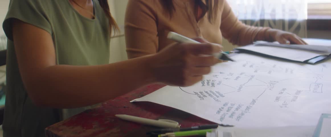 Two women sit at a small table talking and writing on a large piece of paper