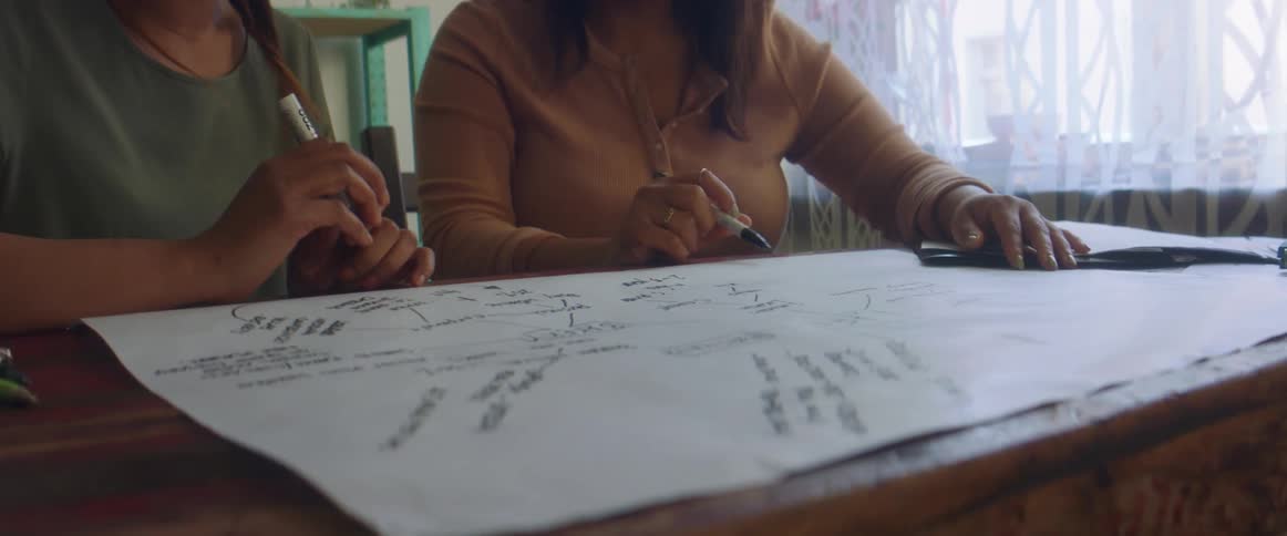 Two women sit at a table in a home and work on a poster