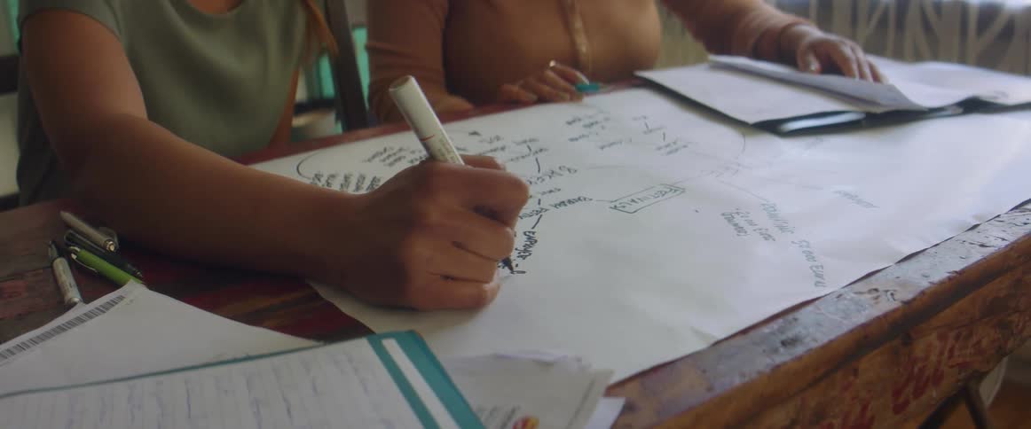 Two women sit at wooden desk writing on paper in black ink as they have discussion