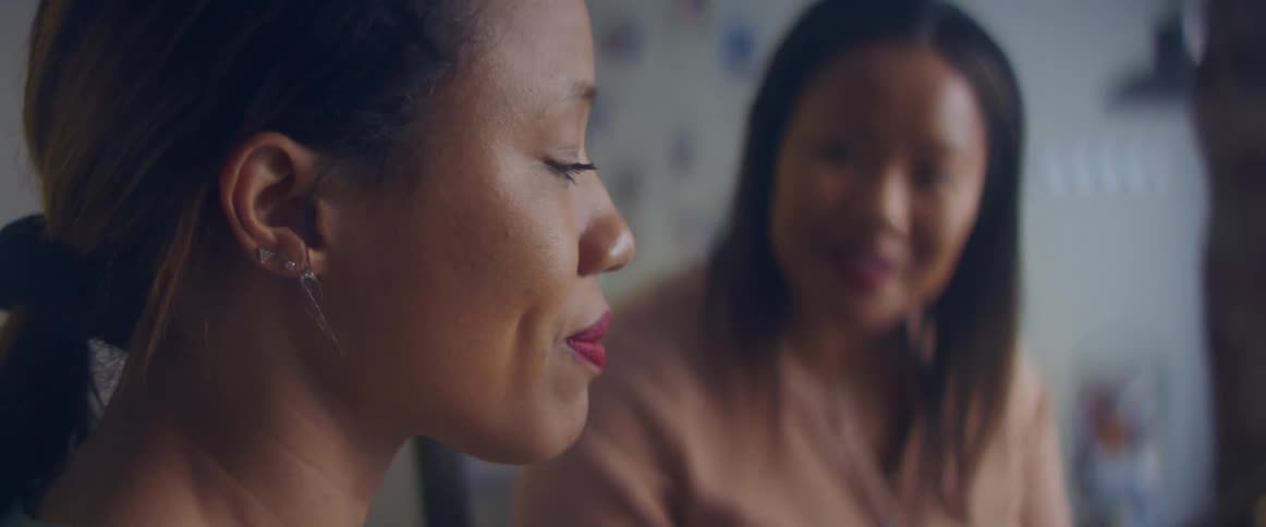 Two women in a room talking with a lamp a wall and a shelf in the background
