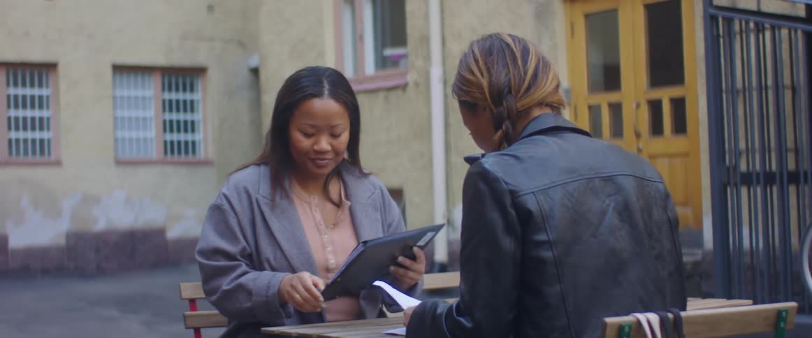 A woman sitting on a chair at a table as another woman walks up and sits down with a building with doors in the background