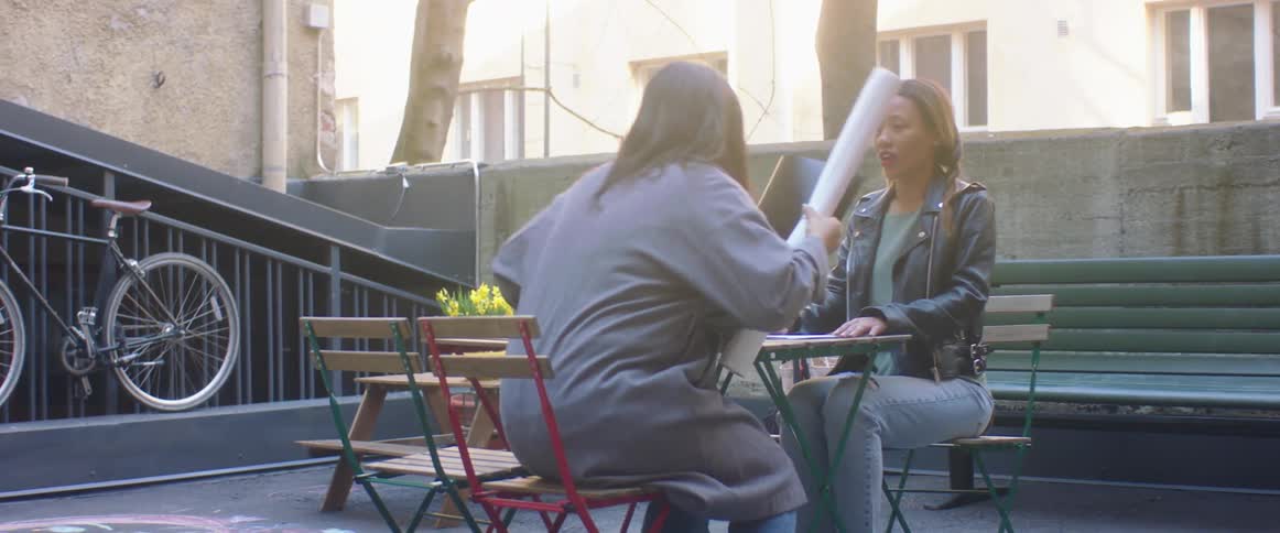 A woman sitting on a chair at a table as another woman walks up and sits down with a bench and a building in the background