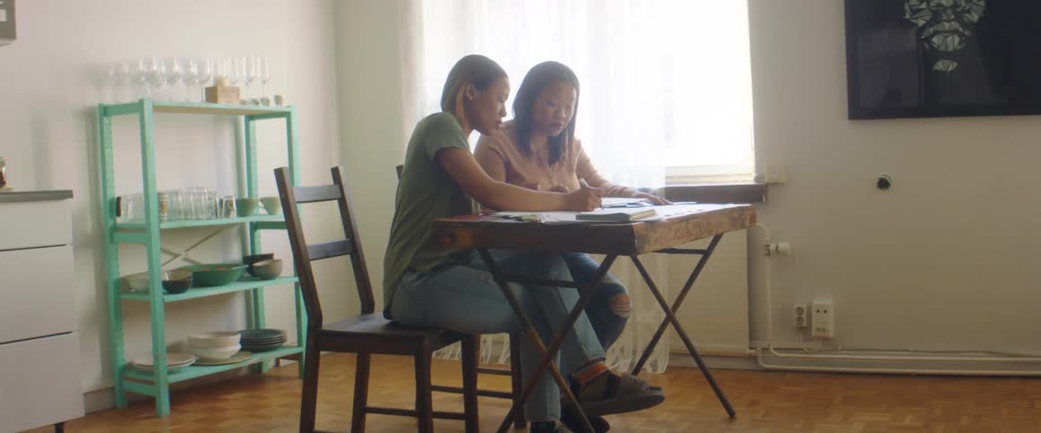 Two women sit together at desk writing on paper as they talk
