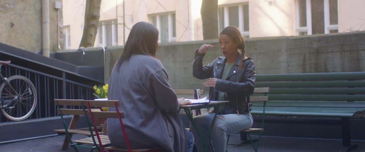 Two woman sitting on chairs at a table with a bench and a wall in the background