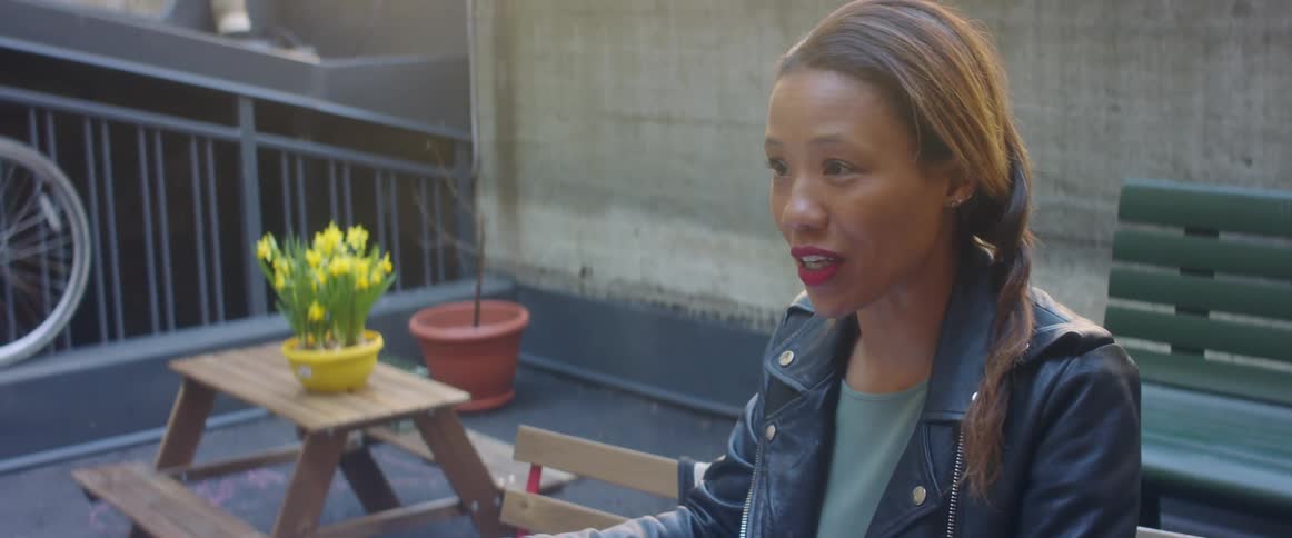A woman sitting on a chair with a table a bench and a wall in the background