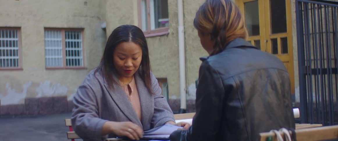 A woman sits at an outdoor table and shows the contents of a binder to another woman sitting across from her