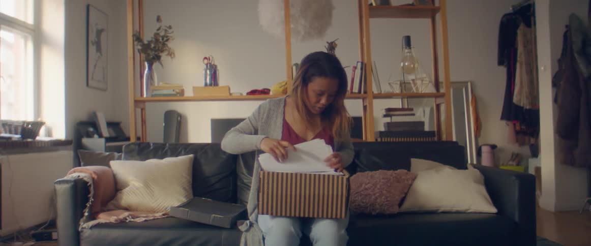 A woman sits down on her couch with a box and rummages through papers inside