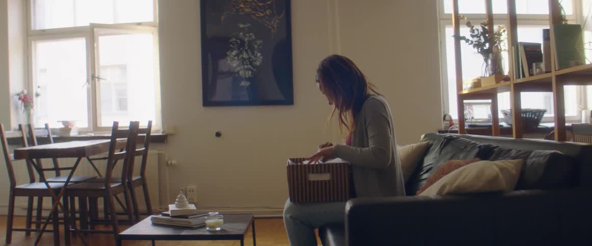 Woman carries a box of old documents into the living room and seats on the couch to check the contents