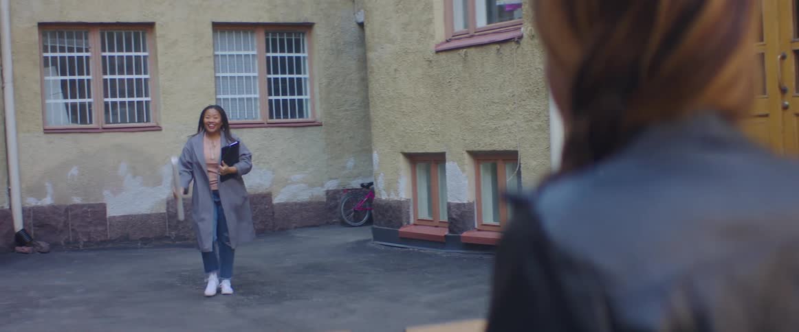A woman carrying a binder and paper walks out of an alleyway towards a waiting woman