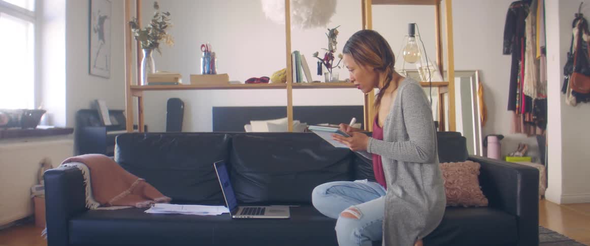A woman sits on a home couch looking at a laptop and writing in a notebook