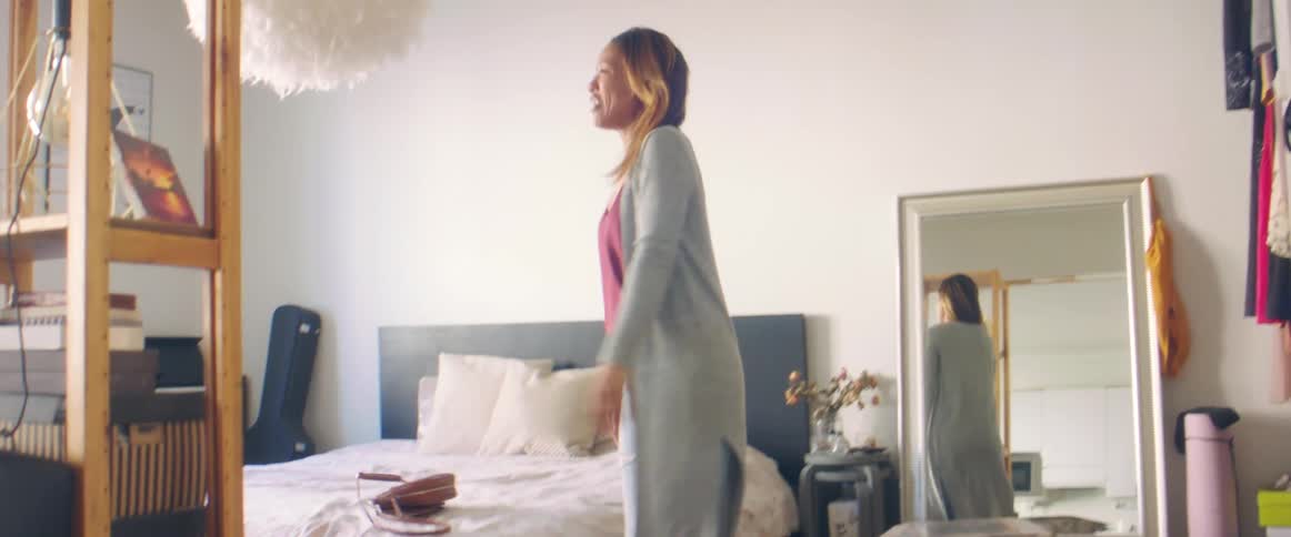 Woman walks into bedroom dances a little before collapsing on bed bordered by guitar case and mirror